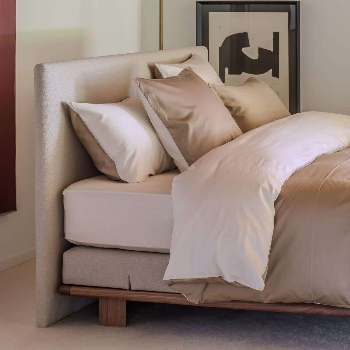 Een bed met een gestoffeerd hoofdbord en lichte satijnen kussens op een houten onderstel